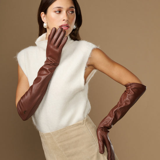 Women’s elbow-length brown leather gloves, one raised near face while wearing.